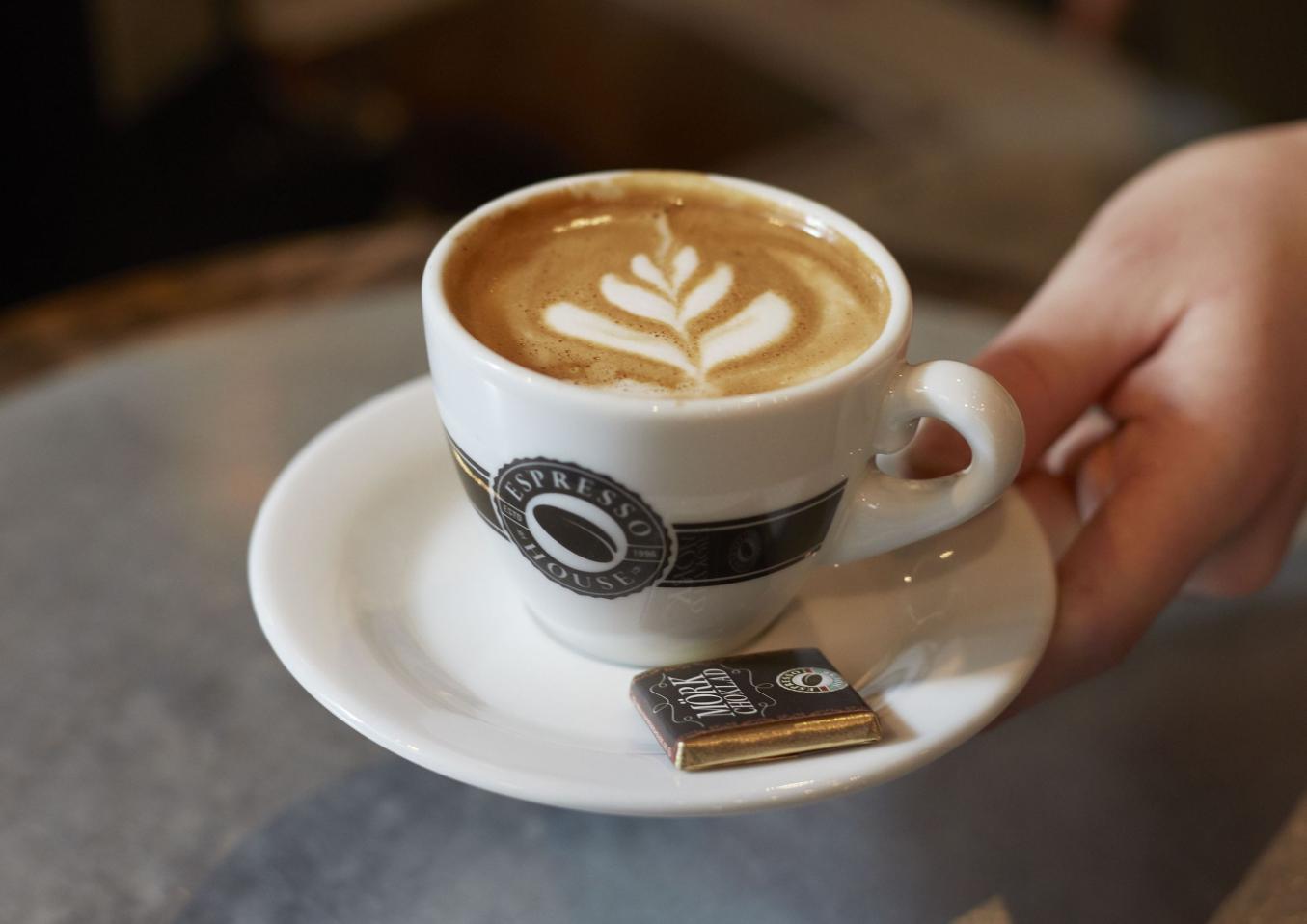 Kuvassa naisen kädessä maitokahviannos valkoisessa kupissa, jonka reunassa Espresso Housen logo. Asetilla ruskeassa kääreessä suklaapala. Kuva lainattu netistä.